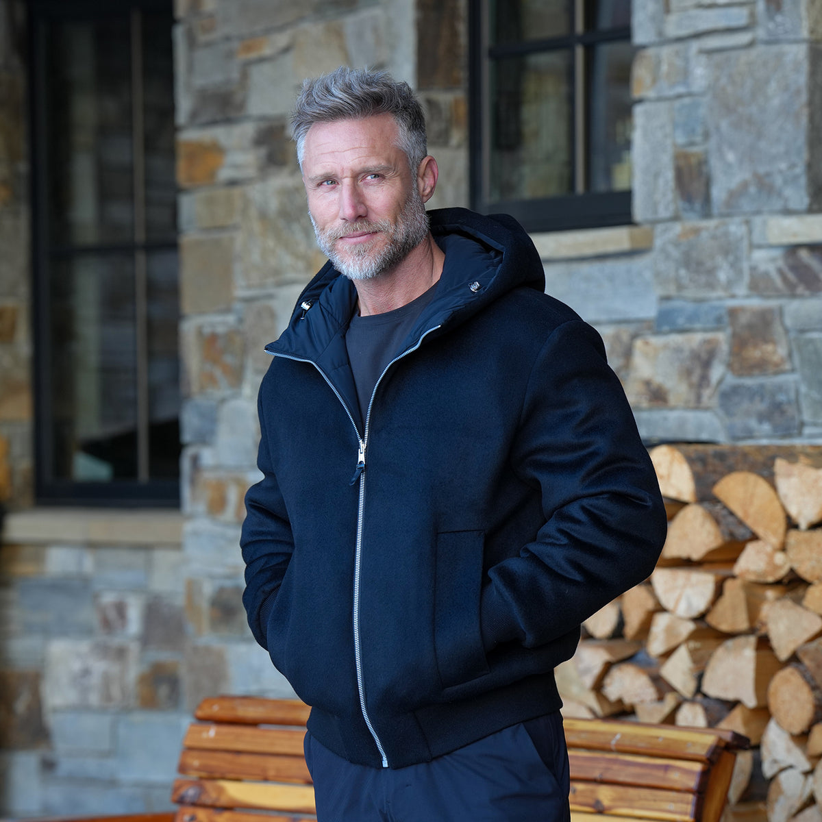 Reversible Wool Puffer in black, hood down, hands in pockets, beside a stacked woodpile