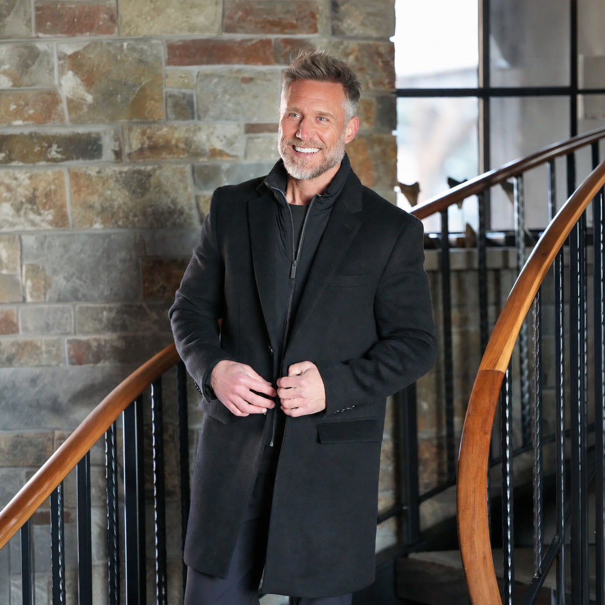 Titan Wool Trench Coat in black, worn open over a zip layer, on a stone staircase